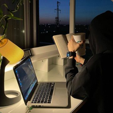 A woman wearing a hijab sits at a desk working on a laptop, with a website open on the screen and a sunset visible through the window in the background.